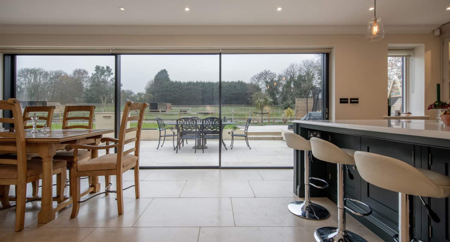Modern kitchen and dining area with wooden table and chairs, bar stools at an island, and large bifold doors opening to a patio with outdoor furniture and a view of a spacious green yard.