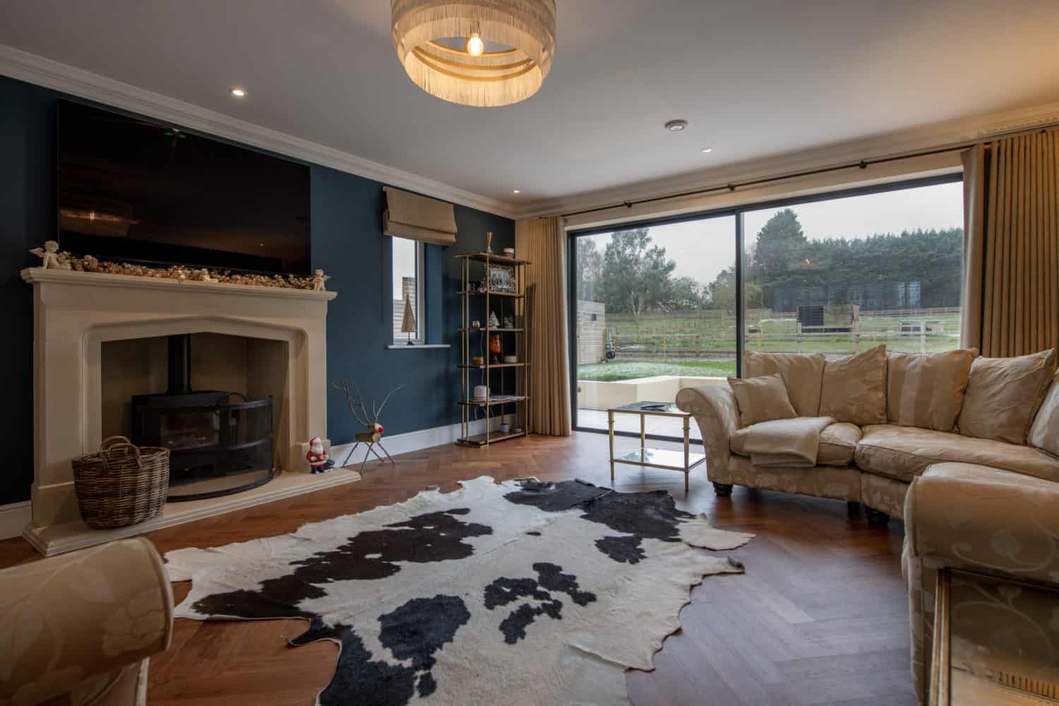 A cozy living room with a curved beige sofa, cowhide rug, fireplace with a wood-burning stove, large TV above the mantel, shelves, and bespoke sliding doors overlooking a garden and trees.