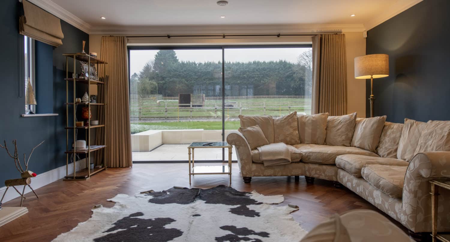 A modern living room with a beige sectional sofa, floor lamp, glass coffee table, and cowhide rug on wooden floors. Large sliding bifold doors offer a view of a green garden and trees outside.