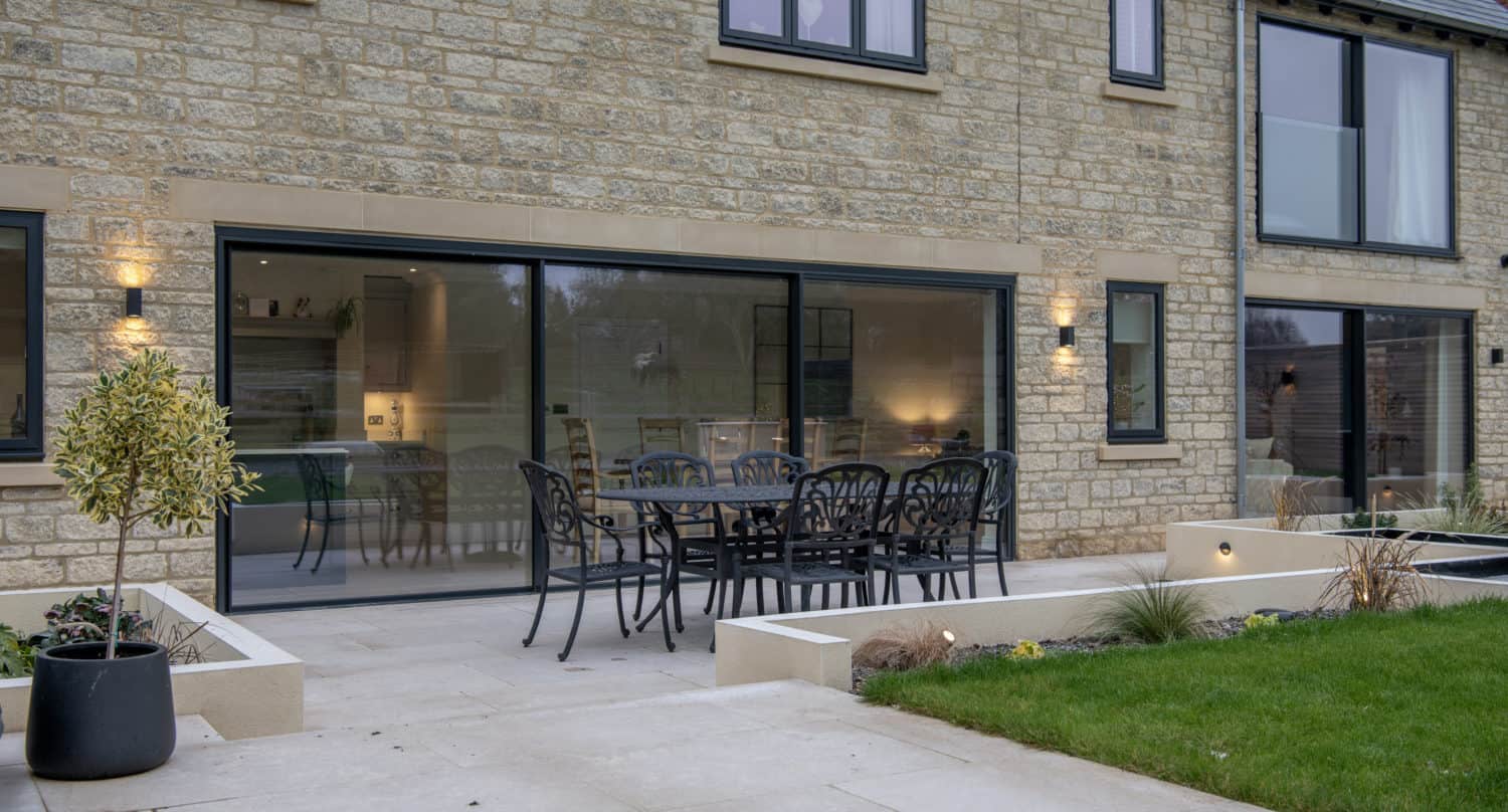 A modern stone house with bifold doors opens to a patio featuring a black outdoor dining set, potted plants, and a small lawn with landscaped garden beds. Warm indoor lights are visible through the glass.