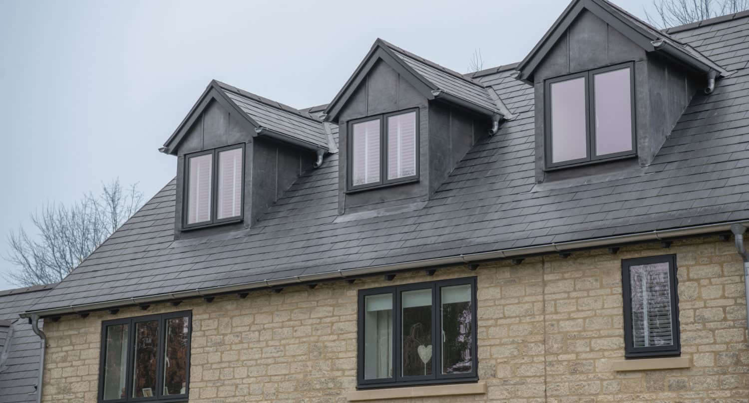 A house with light-colored brick walls and three dark dormer windows on a slate roof features bespoke glass solutions in its front doors. The lower windows reflect outdoor scenery; one window has white blinds and a heart-shaped decoration inside.
