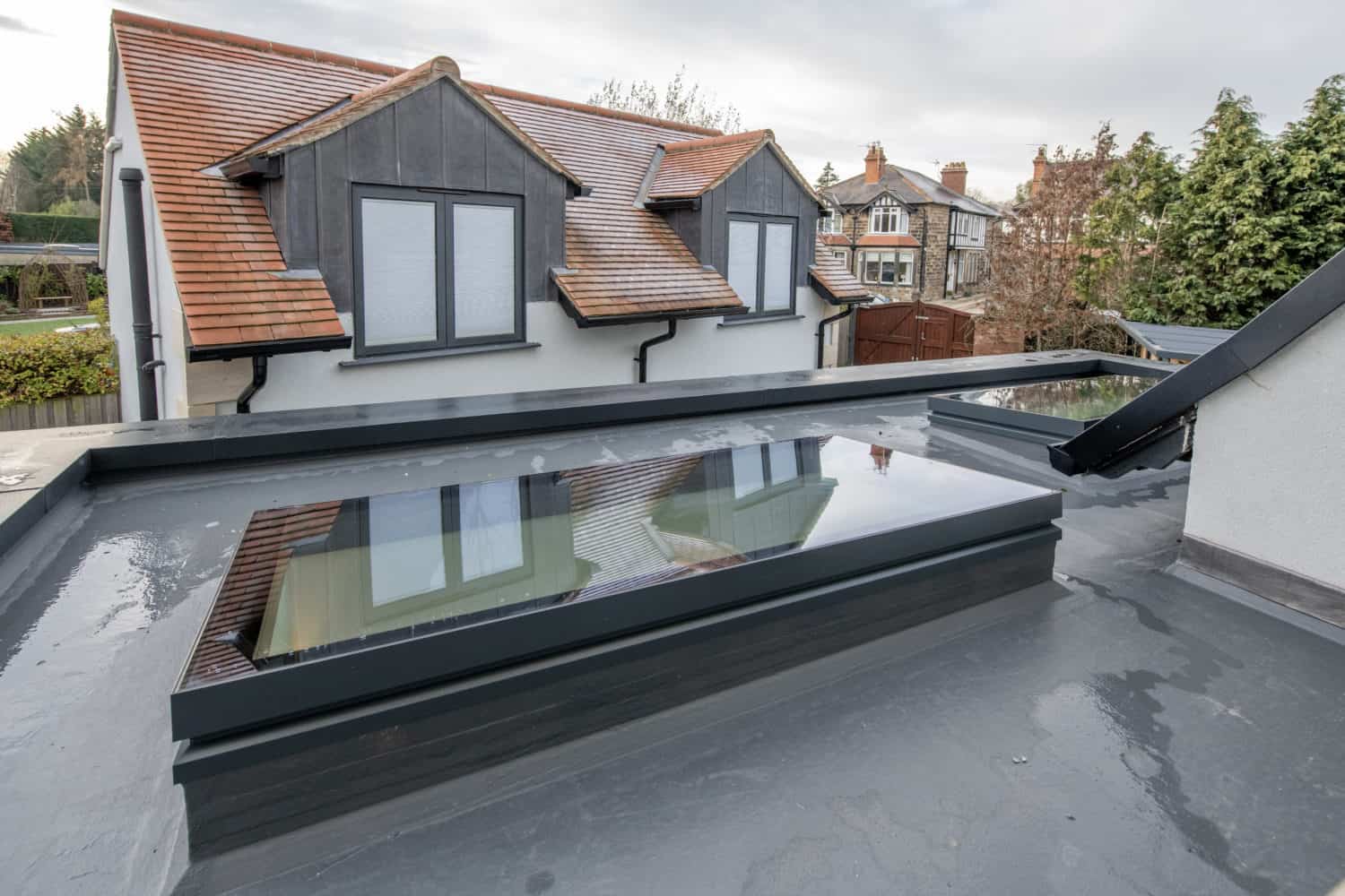 A flat roof with large, rectangular rooflights and standing water, overlooking a house with pitched red-tile roofs, grey-framed windows, and sleek sliding doors on an overcast day.
