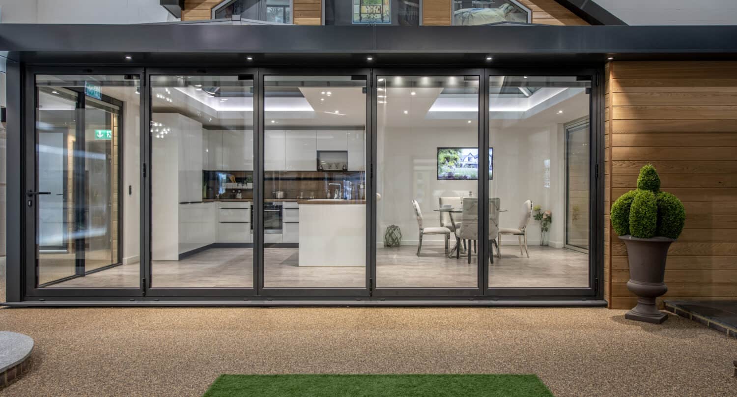 Modern home with bespoke glass solutions, including large bifold doors revealing a bright, open-concept kitchen and dining area. Sleek white cabinets, a dining table, and decorative plants enhance the gravel patio with green artificial grass.