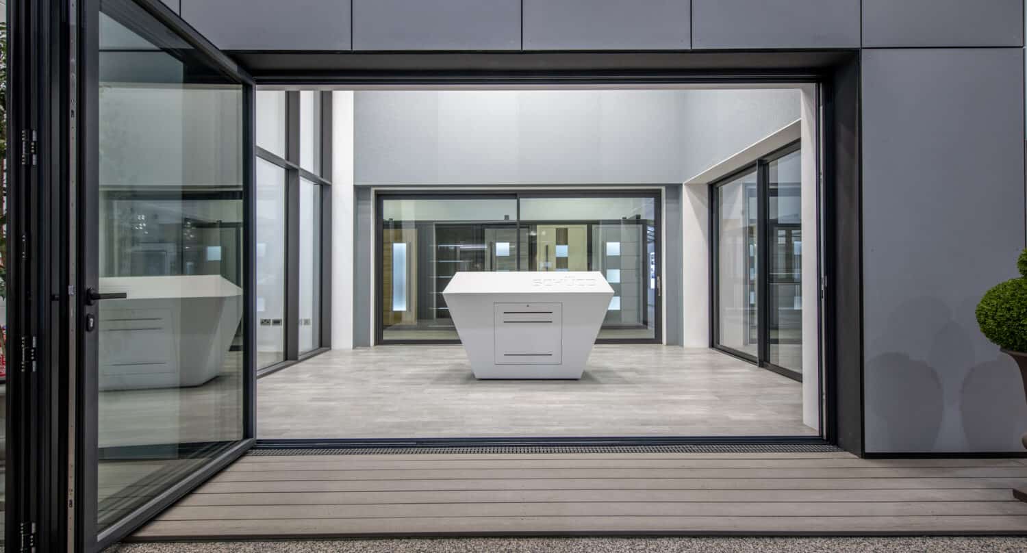 Modern open-concept room with large sliding glass doors, gray flooring, and a central white geometric island. The space is minimalist and bright, featuring clean lines, expansive windows, and a seamless indoor-outdoor flow.