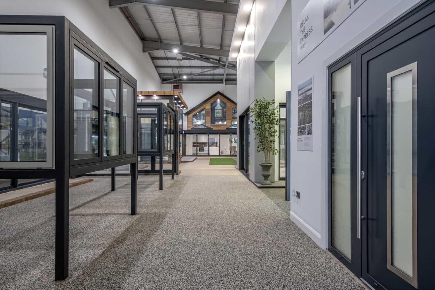 A modern showroom with large windows, front doors, and bifold doors on display, featuring a model house in the background, high ceilings, glass roofing, and bright lighting. The space has a clean, organized layout with neutral colors.