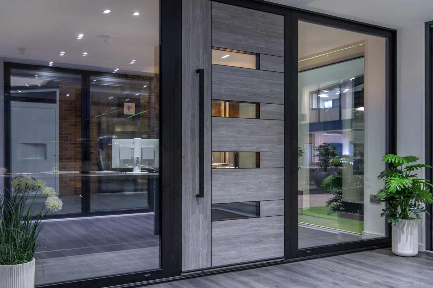 Modern glass and wood front door with horizontal panels and a large black handle, flanked by glass side panels. Indoor plants add freshness, while the space features a contemporary, clean design ideal for stylish front doors.