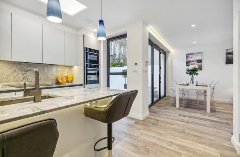 Modern kitchen with marble countertops, built-in appliances, two bar stools, and pendant lights. Open dining area with a table set for four, glass chairs, large windows, and bifold doors letting in natural light.