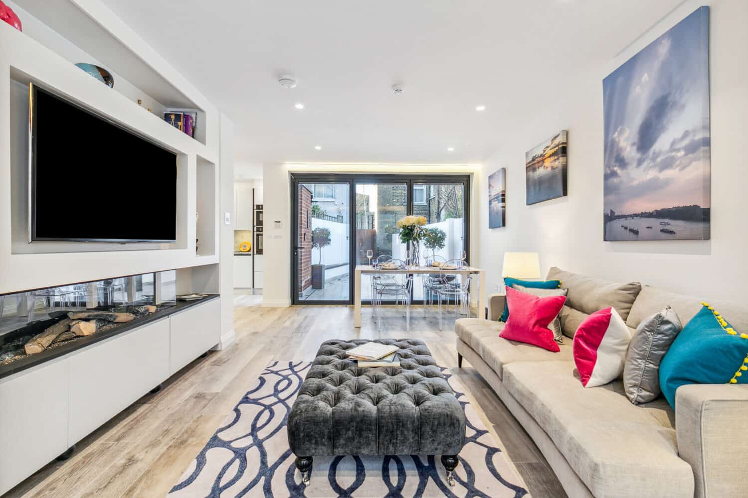 Modern living room with a beige sofa, colorful pillows, and a tufted ottoman. Wall-mounted TV, fireplace, and artwork. Bifold doors open to a patio, and a dining table with chairs is visible in the background.