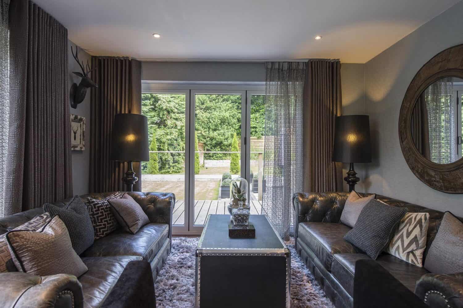 A modern living room with two dark leather sofas, patterned cushions, a trunk-style coffee table, two black lamps, a textured rug, and large sliding doors offering bespoke glass solutions overlooking a green garden.
