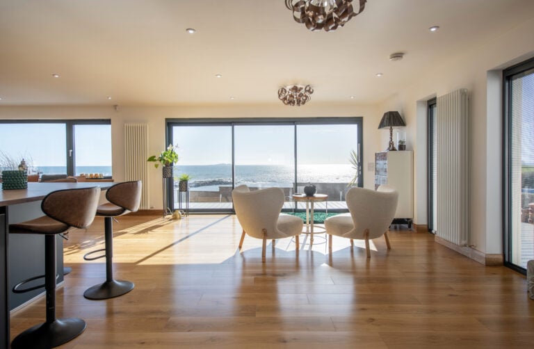 A bright, modern living space with large windows and bifold doors overlooking the ocean features wooden floors, two cozy chairs around a small table, and bar stools at a counter. Sunlight floods the room, creating a warm and inviting atmosphere.