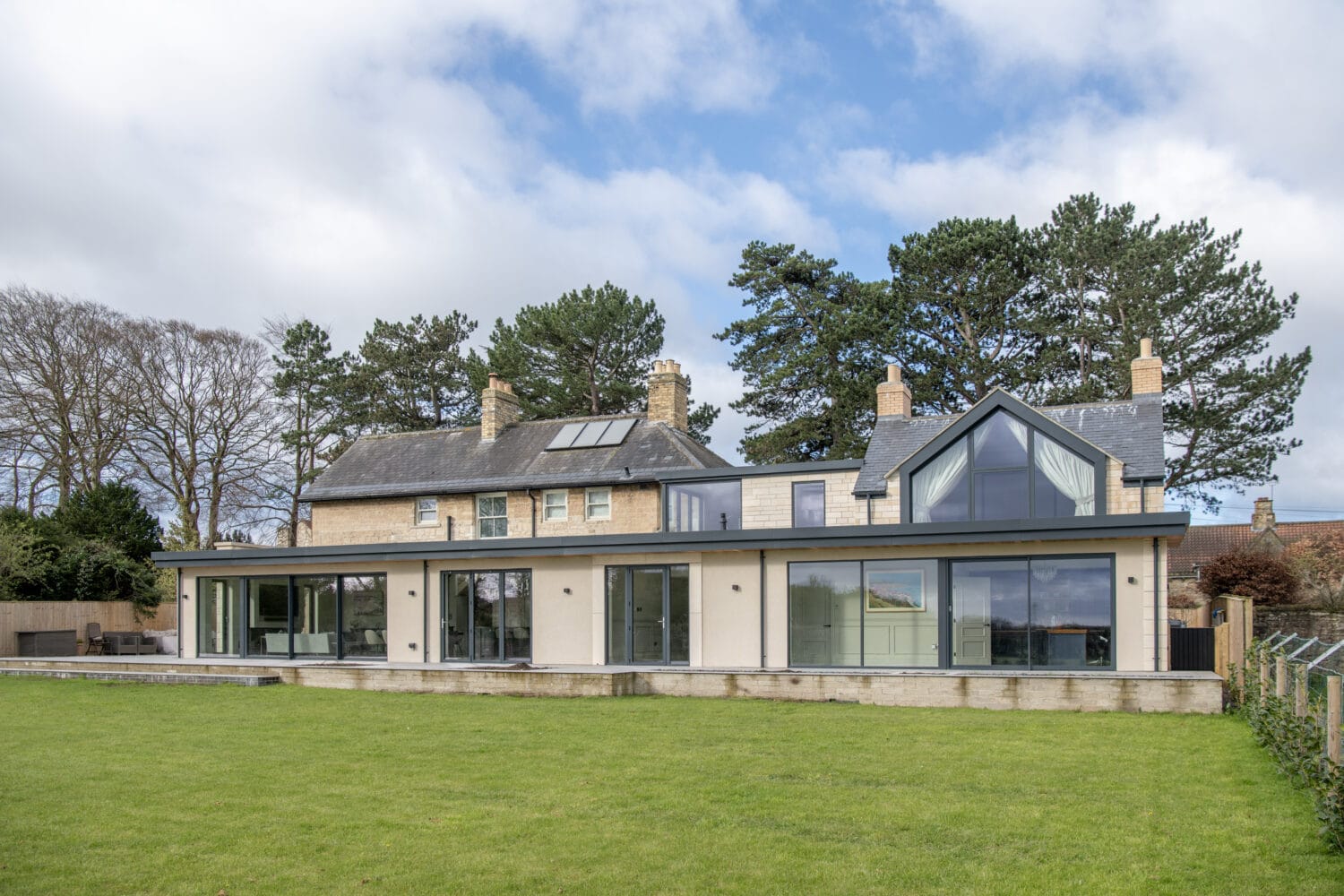 A large modern house with stone and glass features, tall windows, bifold doors, multiple chimneys, and a spacious green lawn in front, set against a backdrop of trees and a partly cloudy sky.