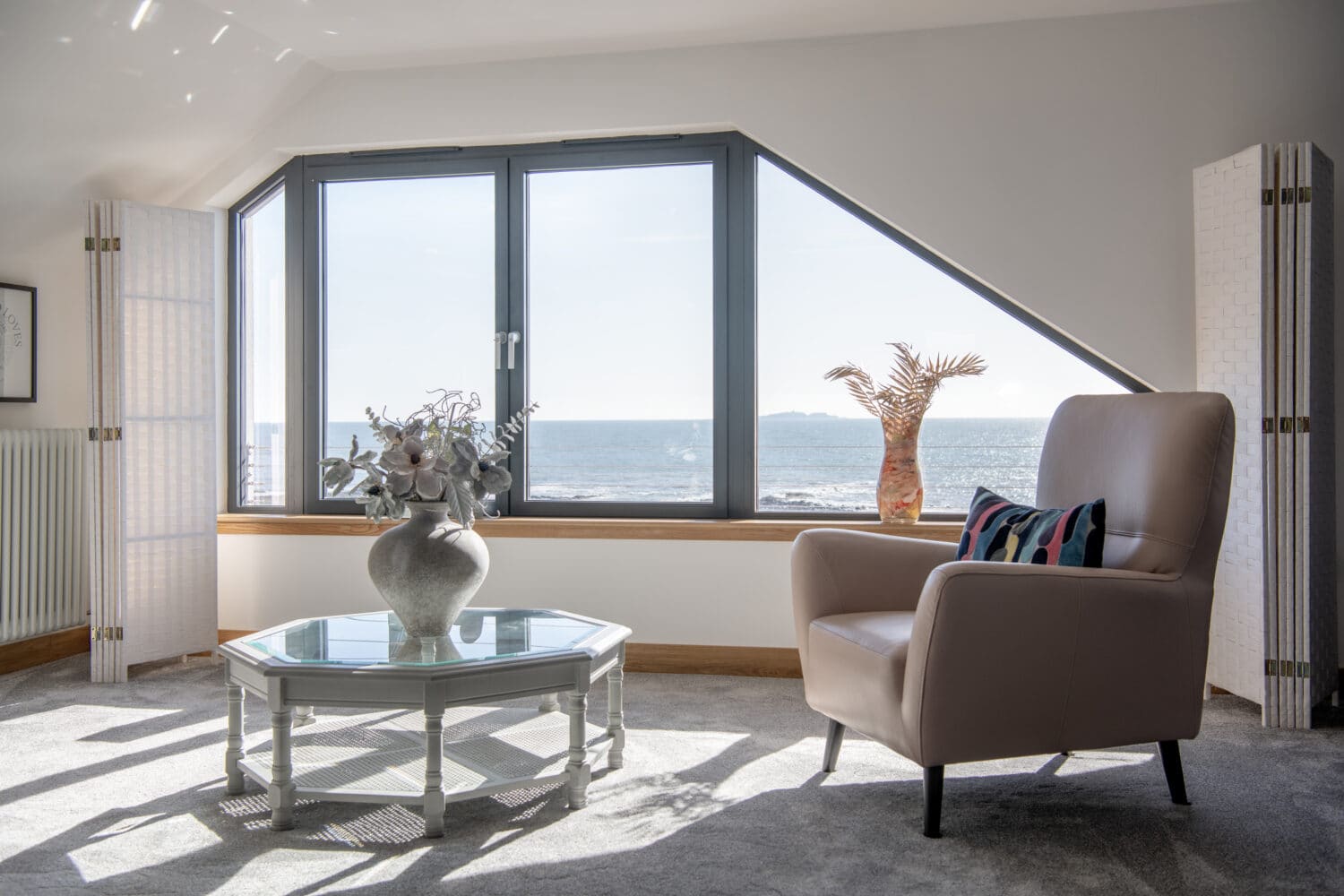 A bright coastal living room with a large window overlooking the sea, a beige armchair, glass coffee table with a vase, and a decorative palm plant by the new bifold doors. Sunlight fills the space.