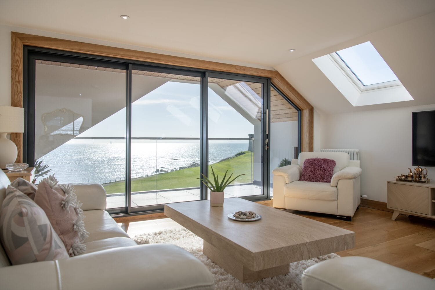 Modern living room with large bifold doors opening to a balcony overlooking the ocean, bright natural light, cozy armchairs, a beige coffee table, and wooden floors with a plush white rug.
