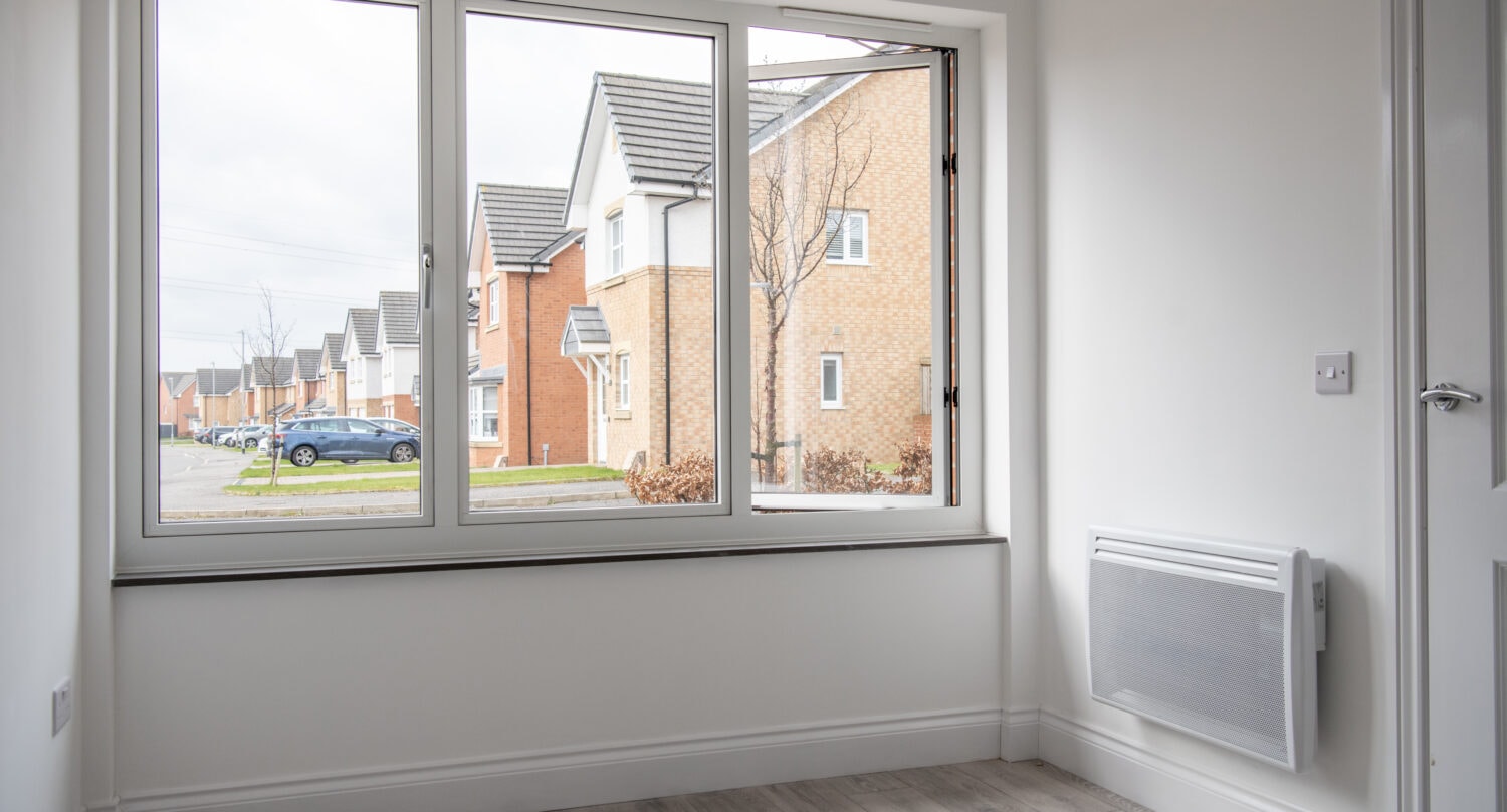 A bright, empty room with white walls and light wood flooring, featuring a large window with bespoke glass solutions that looks out onto a suburban street lined with modern brick houses and parked cars.