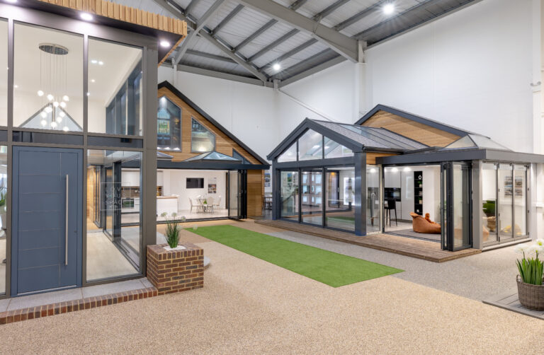 A modern indoor showroom displays two glass-walled home extensions with pitched roofs, stylish lighting, lounge furniture, bifold doors, and artificial grass set within a large, bright industrial-style space.