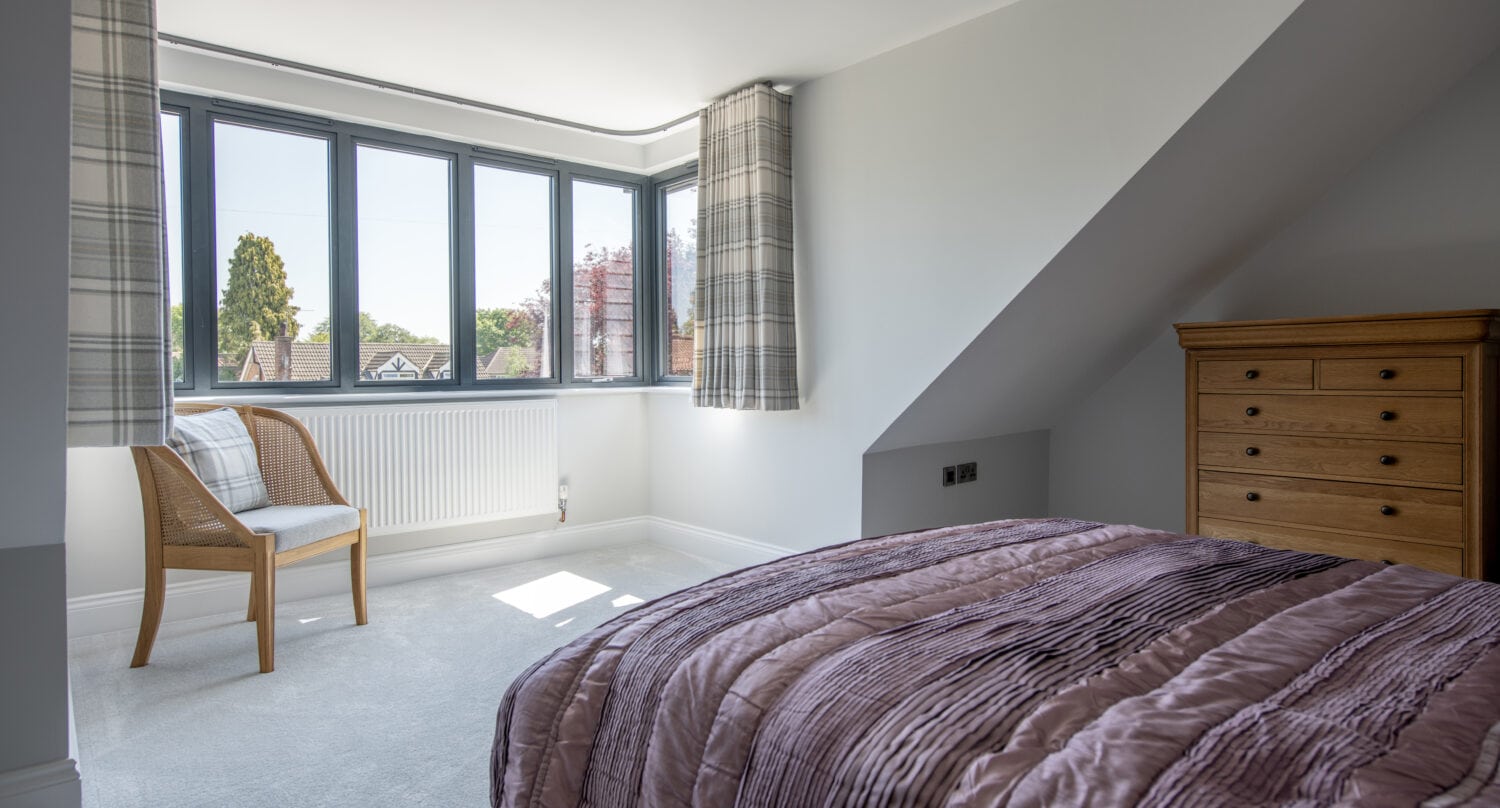 A bright bedroom with a large window, light gray walls, a wooden chair with a plaid cushion, and a bed with a mauve quilted blanket. Sunlight streams in through bifold doors, creating a cozy atmosphere.