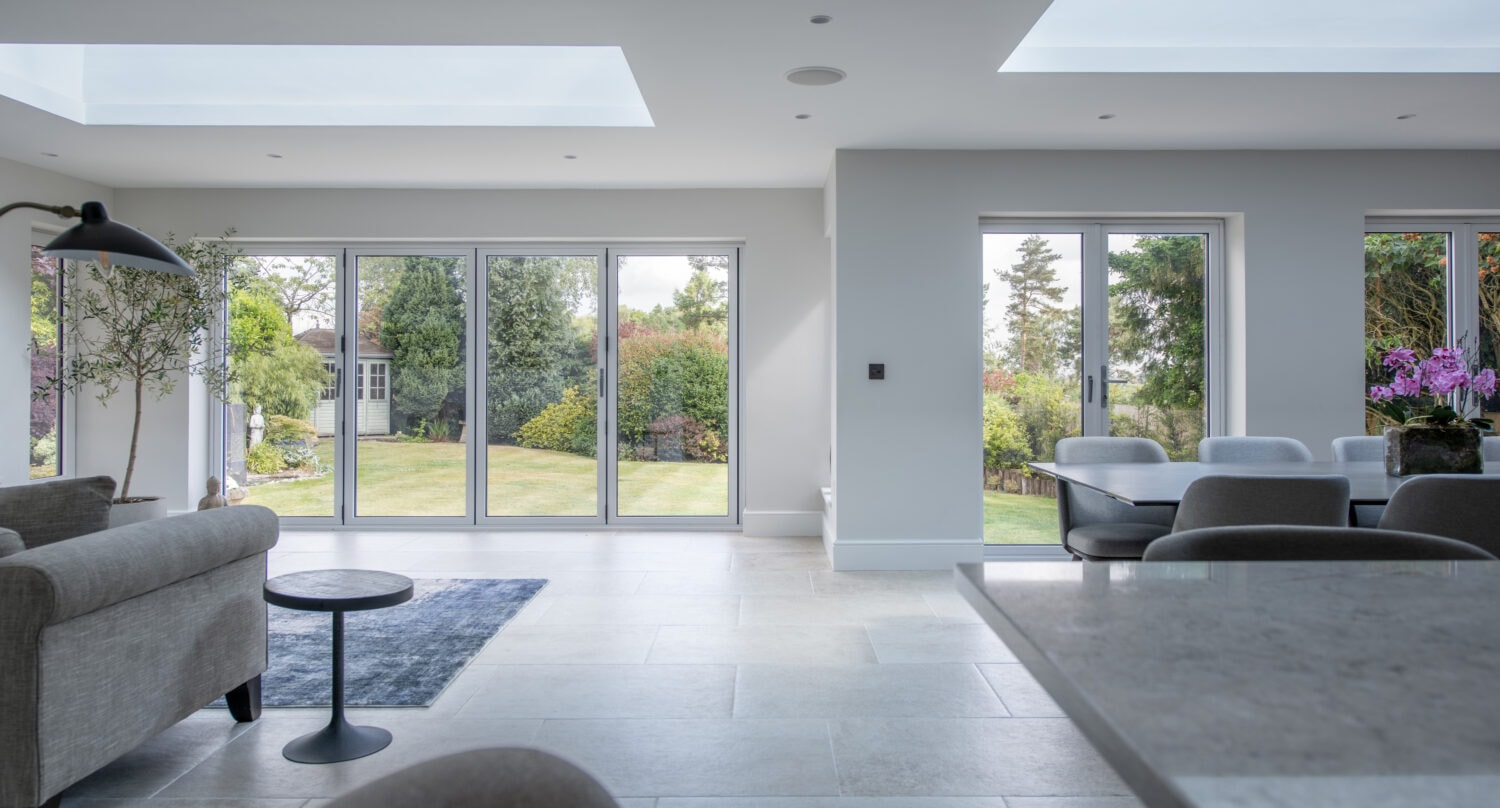 Modern, bright living and dining area with large bifold doors opening to a garden. Light gray furniture, a dark rug, skylights, and a dining table with flowers create an airy, inviting atmosphere.