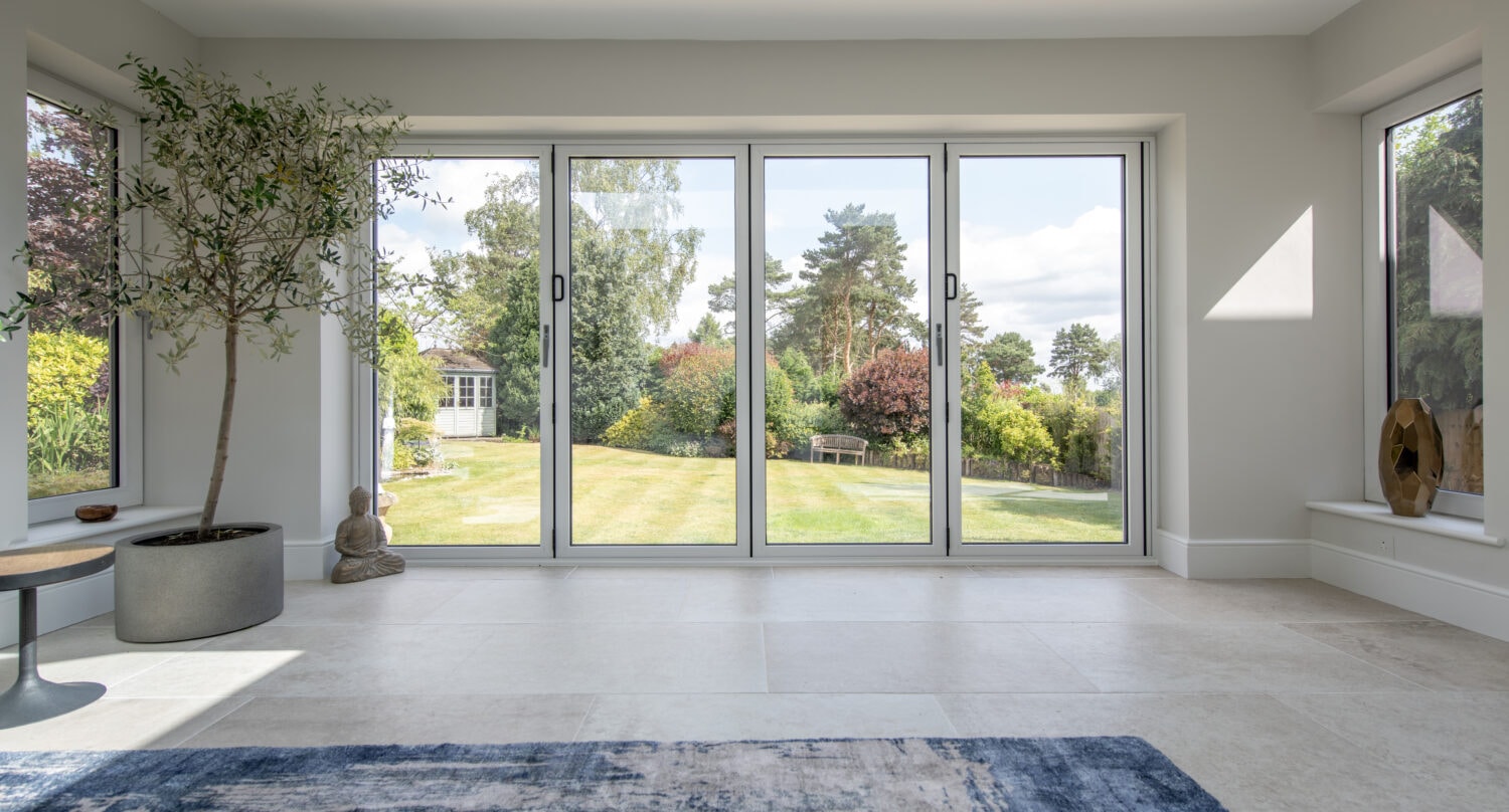 A bright, modern room with large bifold doors overlooking a spacious garden. Indoor plants, a small table with a Buddha statue, and a blue rug decorate the light-tiled floor. Trees and greenery are visible outside.