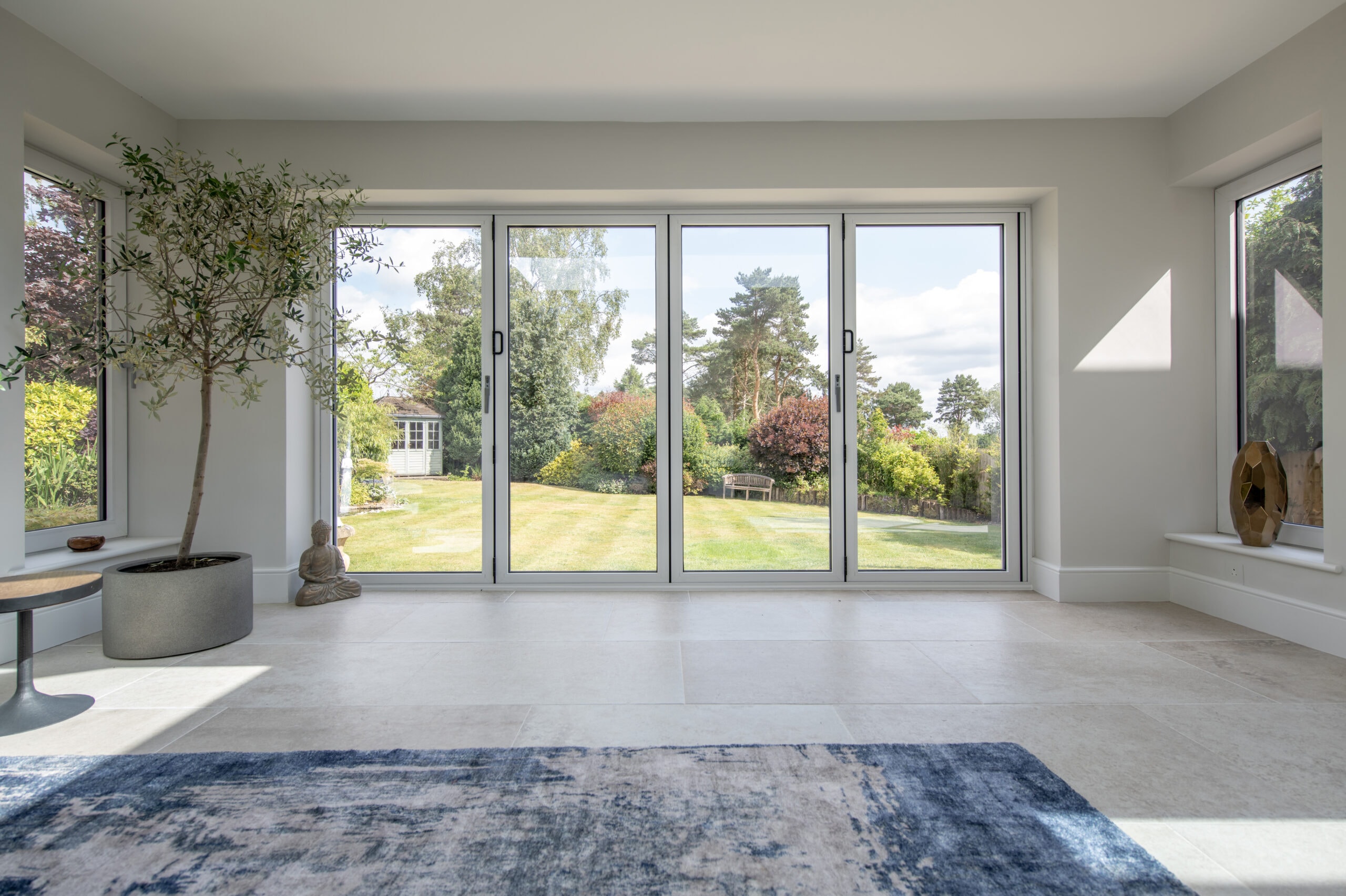 A bright, modern room with large bifold doors overlooking a spacious garden. Indoor plants, a small table with a Buddha statue, and a blue rug decorate the light-tiled floor. Trees and greenery are visible outside.