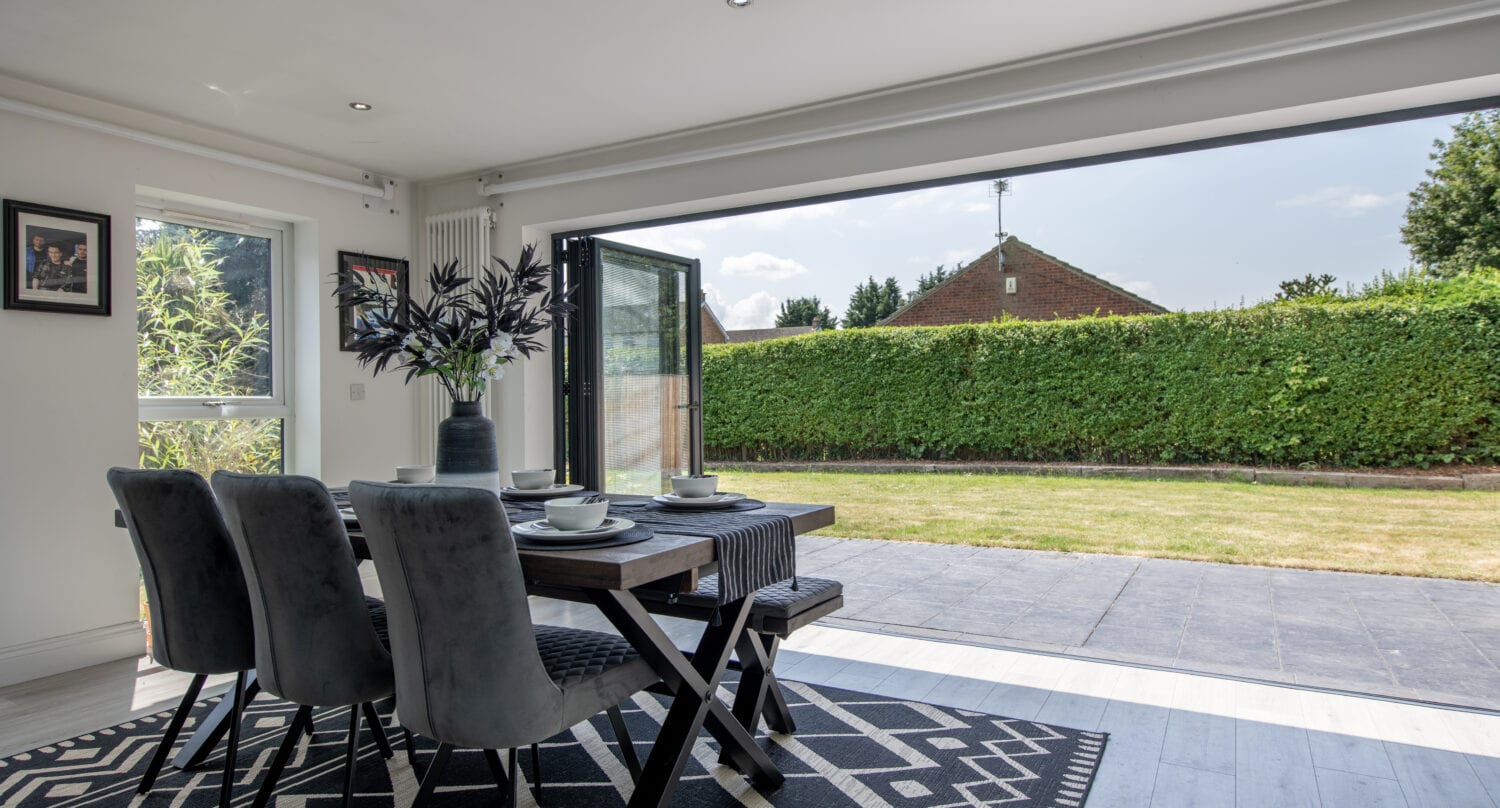 A modern dining area with a black table and four gray chairs sits on a patterned rug beside wide open sliding doors, overlooking a green garden and a hedged lawn under a sunny sky.
