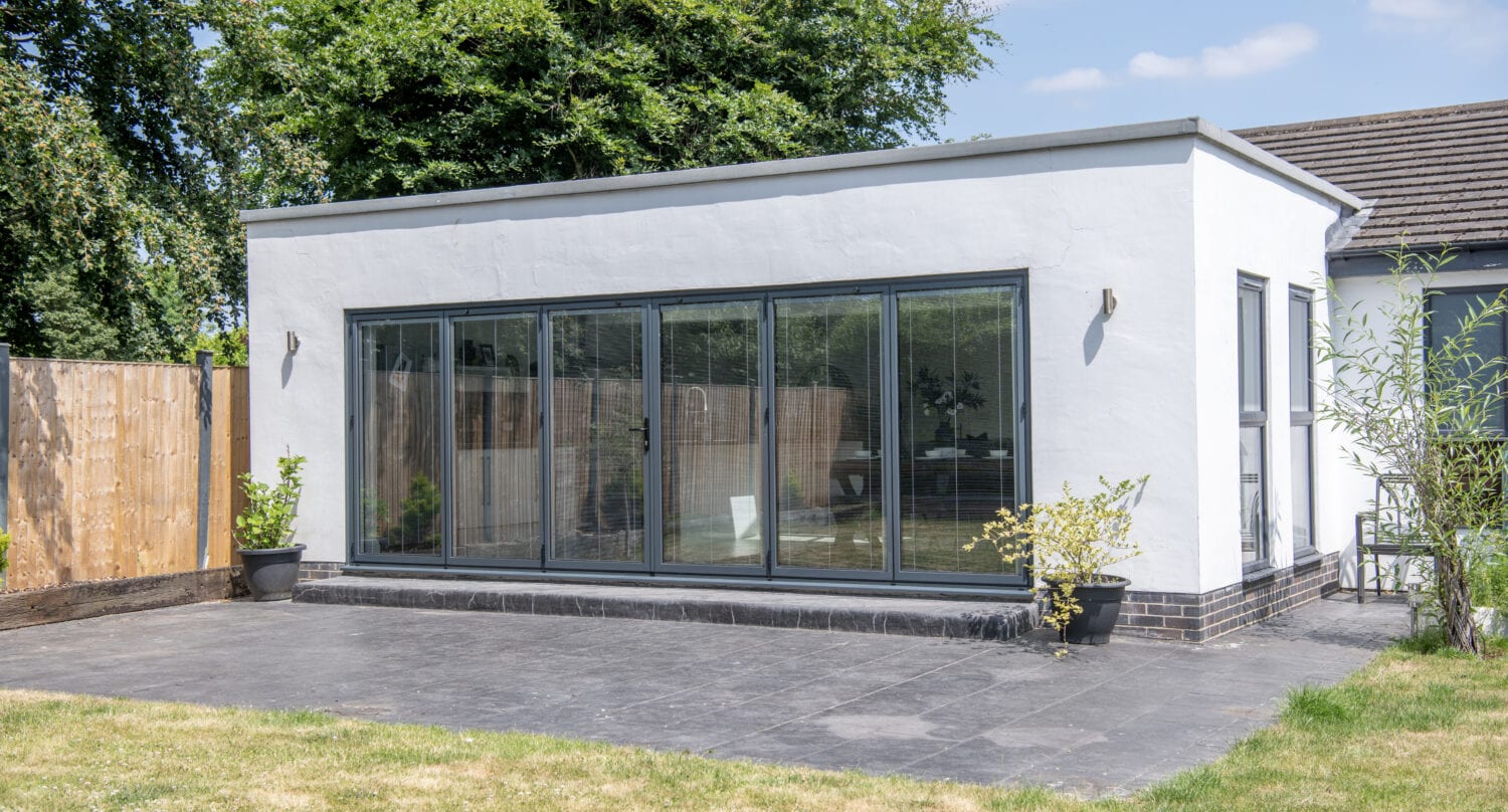 A modern white house extension with large sliding doors, a flat roof, outdoor wall lights, and small potted plants on a tiled patio, surrounded by a grassy yard and wooden fence.