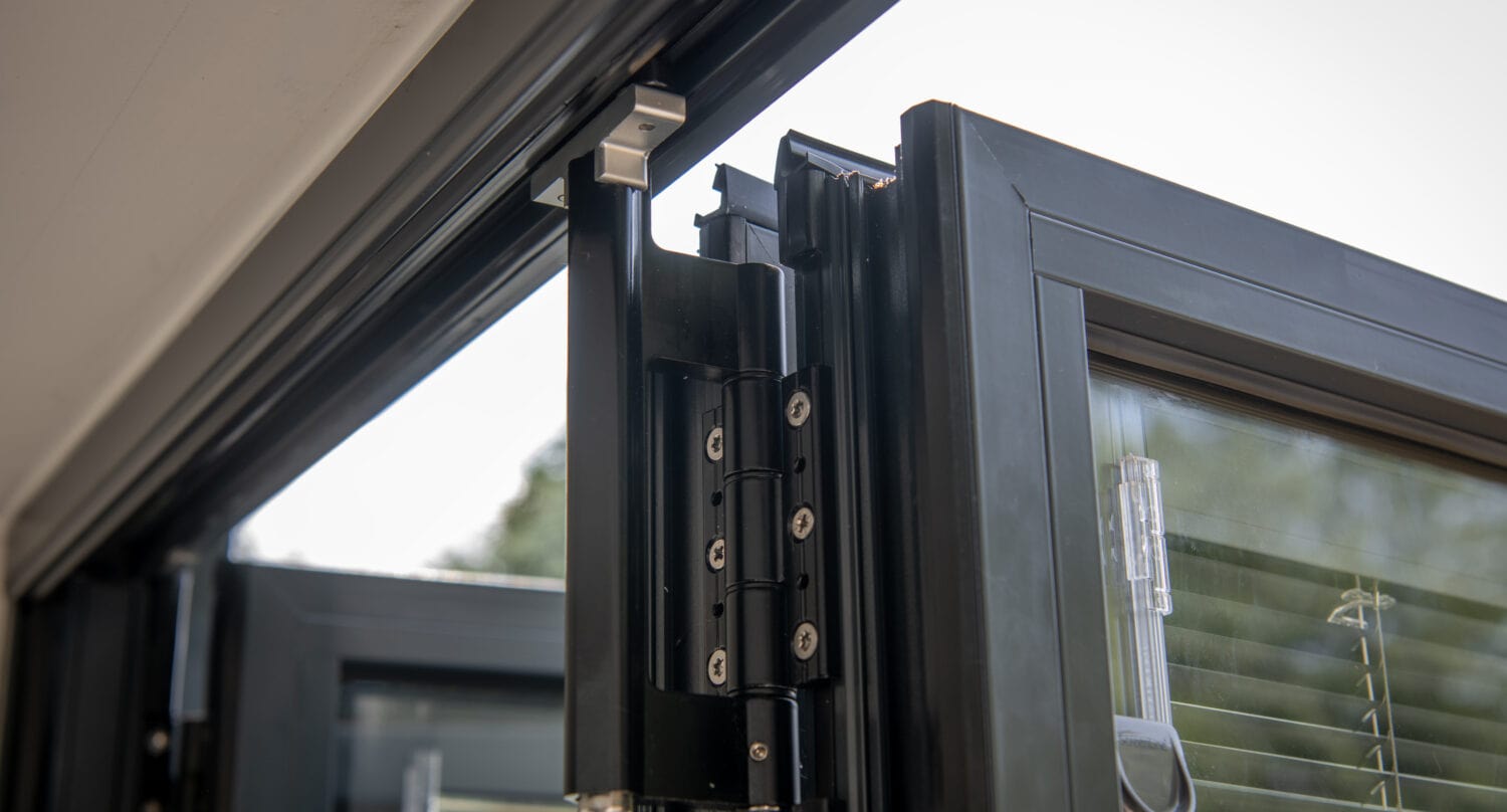 Close-up of a modern black aluminum bi-fold door hinge mechanism, showing the bifold doors partially open with glass panels and integrated blinds visible in the background.