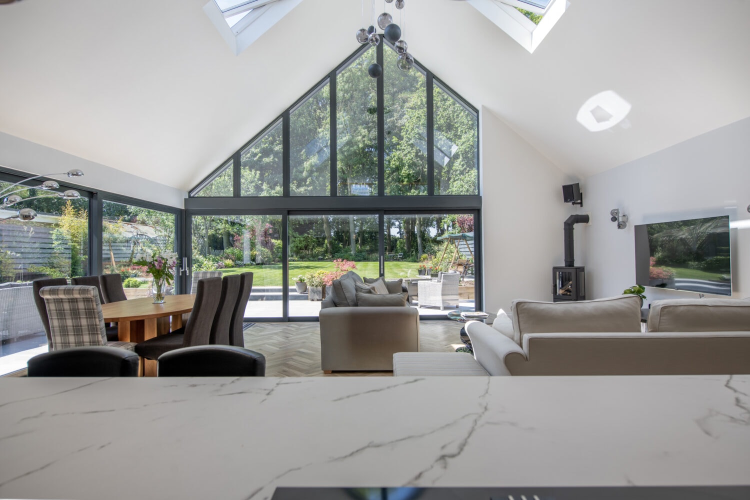 Modern, spacious living room with high vaulted ceiling, large bifold doors and skylights, neutral furniture, dining area with chairs, a garden view, and natural light filling the space.
