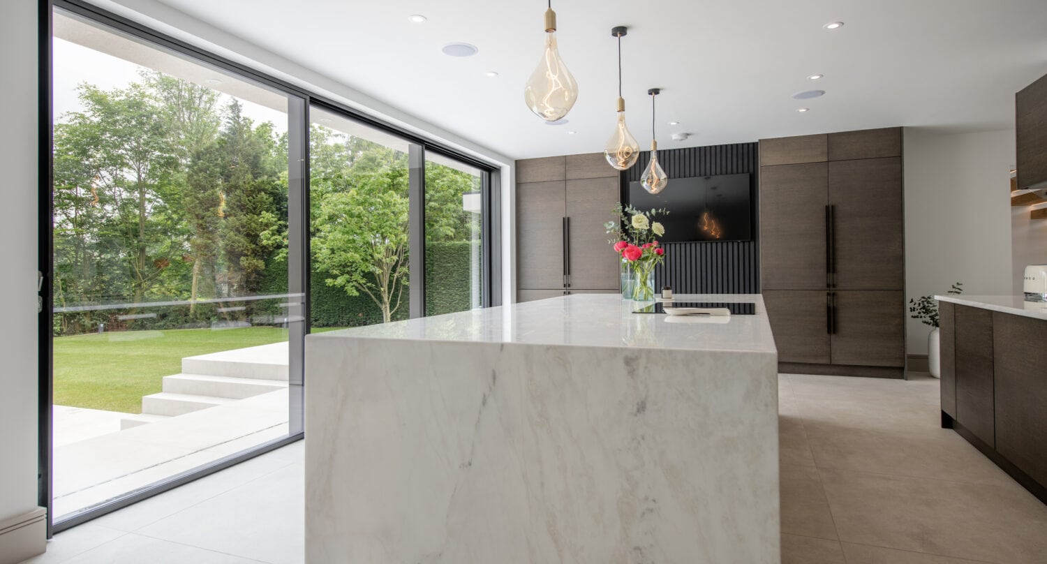 Modern kitchen with a large white marble island, pendant lights, sleek dark cabinetry, and floor-to-ceiling sliding doors opening to a lush green garden. Natural light fills the spacious, minimalist space.
