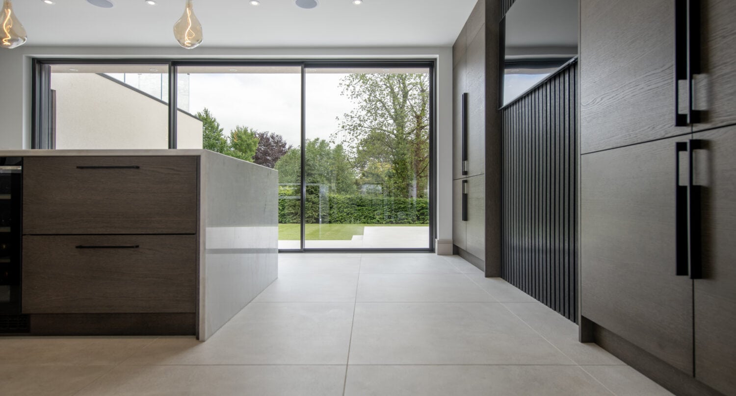 Modern kitchen with large gray floor tiles, wood cabinetry, an island, pendant lights, and sliding doors opening to a green garden with trees and hedges.