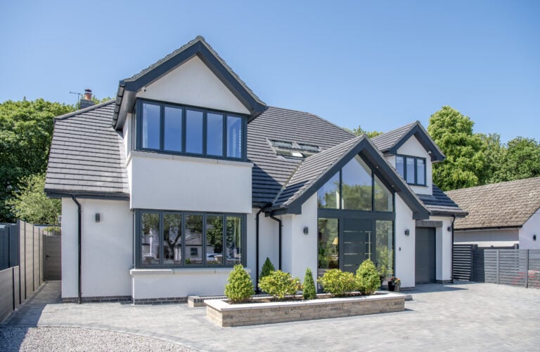 Modern two-story house with large windows, bespoke glass solutions, a gray roof, and white walls. Landscaped bushes line the front, with a paved driveway and trees in the background on a clear, sunny day.