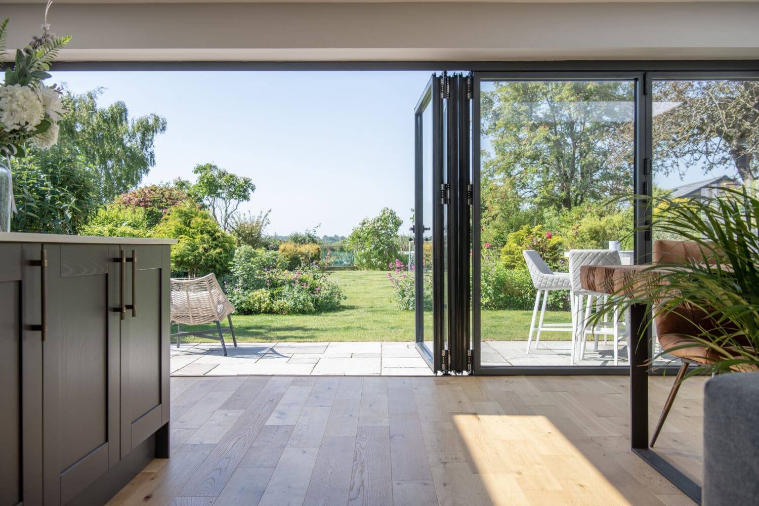 Modern kitchen and dining area with large glass bi-fold doors, complemented by bespoke glass solutions, opens to a sunny garden patio featuring greenery, outdoor seating, and a bright, inviting atmosphere.