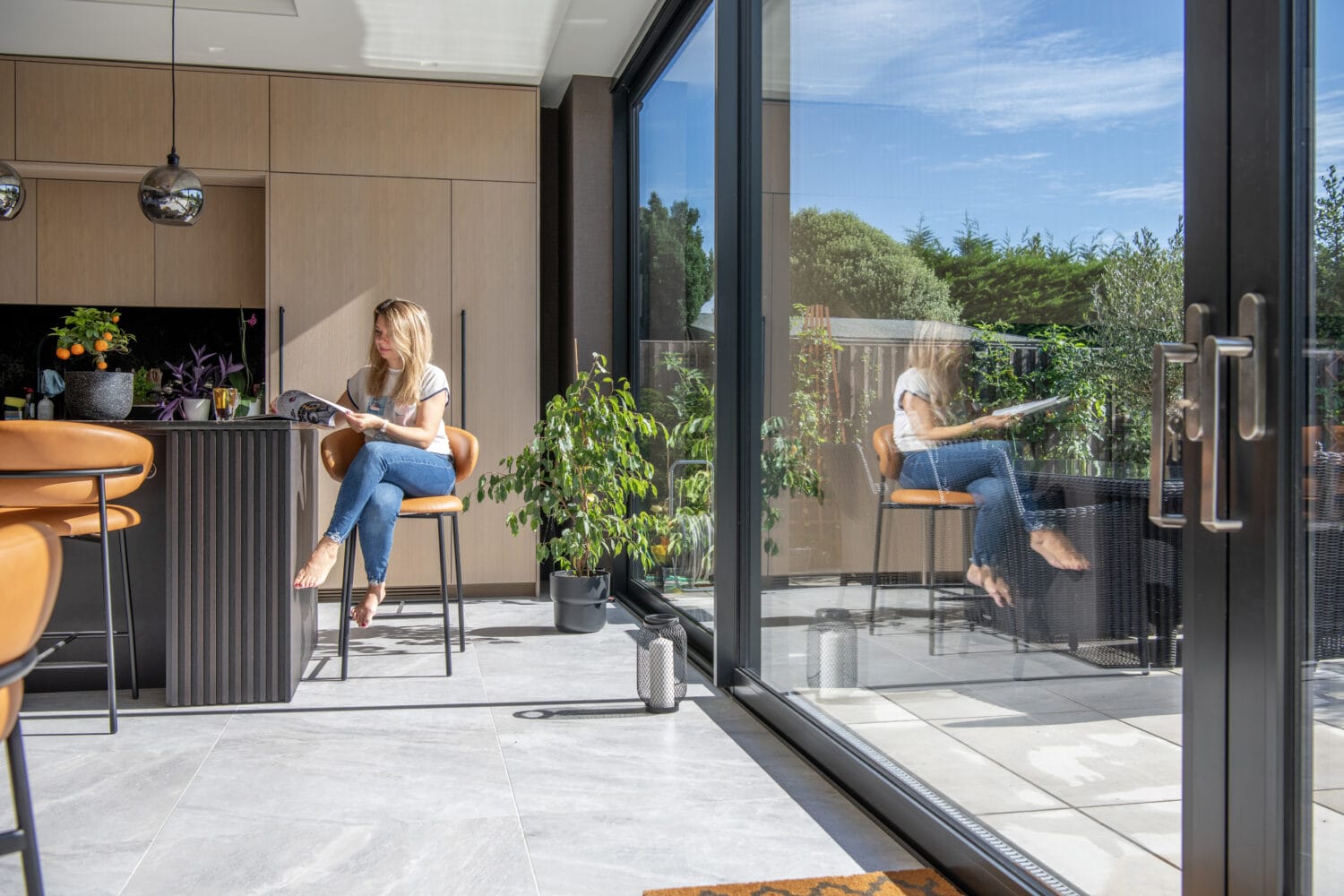A woman sits on a stool in a modern kitchen, reading a magazine. Bespoke glass solutions create large doors that reflect her image, revealing a sunny patio and green garden. The space is bright with natural light and contemporary decor.