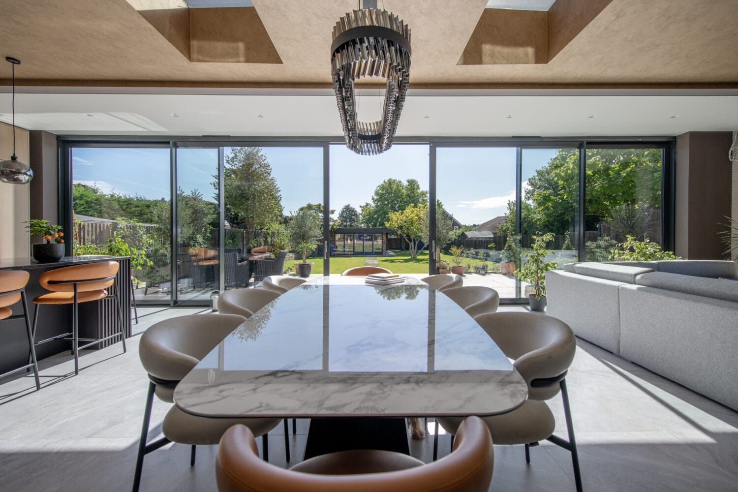 Modern dining room with a marble table, stylish chairs, and large bifold doors opening to a sunny garden. Bar stools line a kitchen island, and a grey sofa sits to the right under skylights.