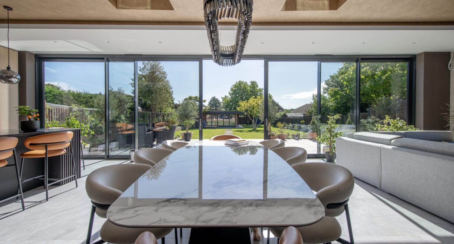 Modern dining room with a marble table, stylish chairs, and large bifold doors opening to a sunny garden. Bar stools line a kitchen island, and a grey sofa sits to the right under skylights.