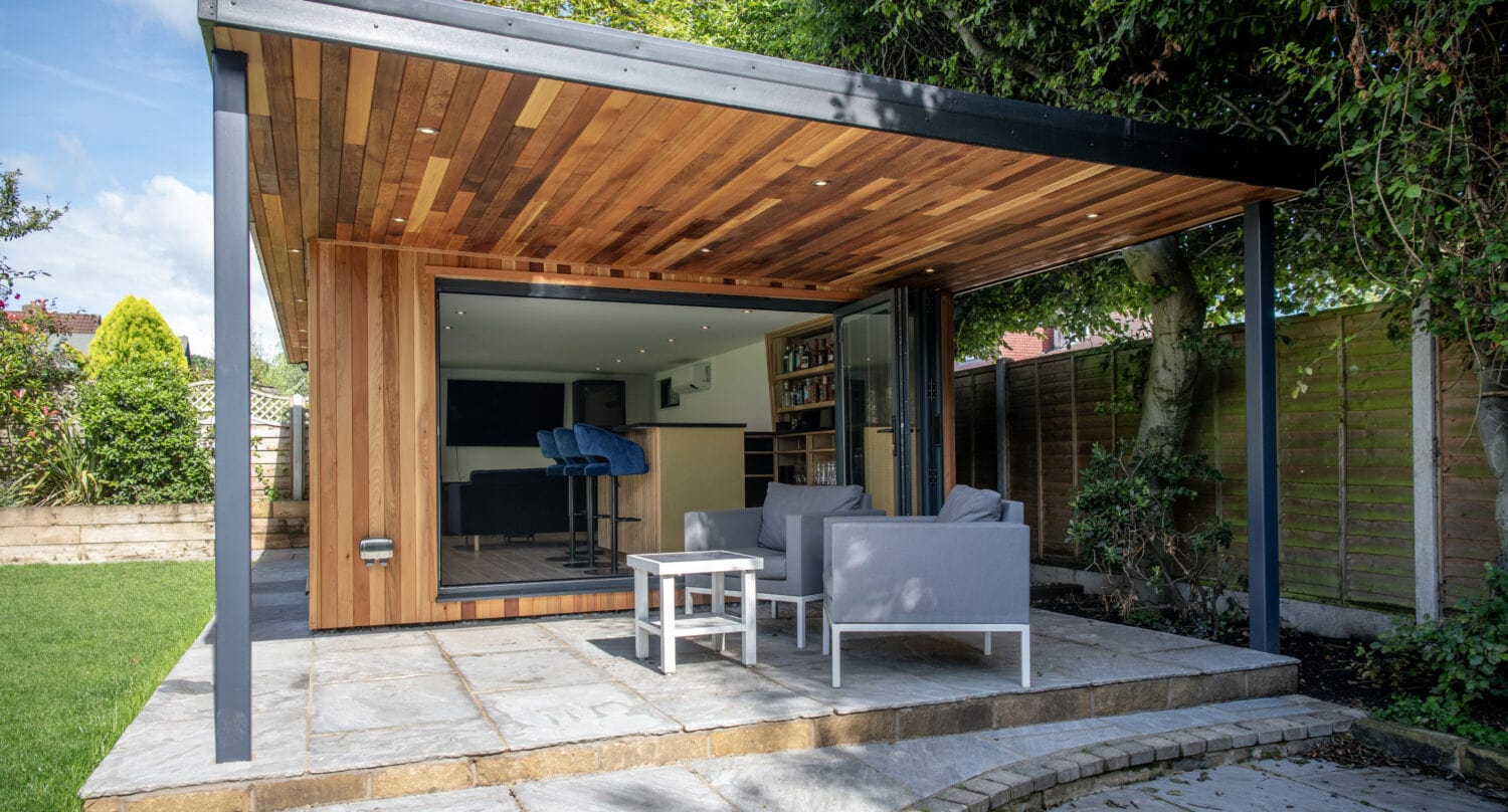 Modern garden room with a wooden ceiling and open bifold doors, featuring outdoor seating on a stone patio, surrounded by greenery and a wooden fence—a showcase of bespoke glass solutions.