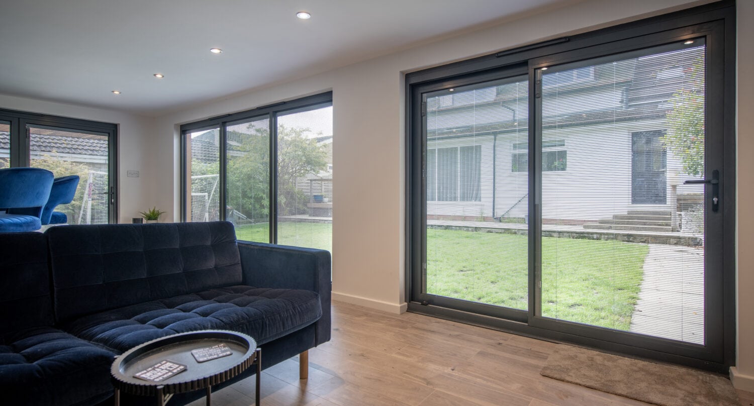 Modern living room with a dark blue sofa, round coffee table, and large sliding glass doors offering a view of a green backyard and neighboring house. Light wood flooring and bespoke glass solutions create a bright, open space.