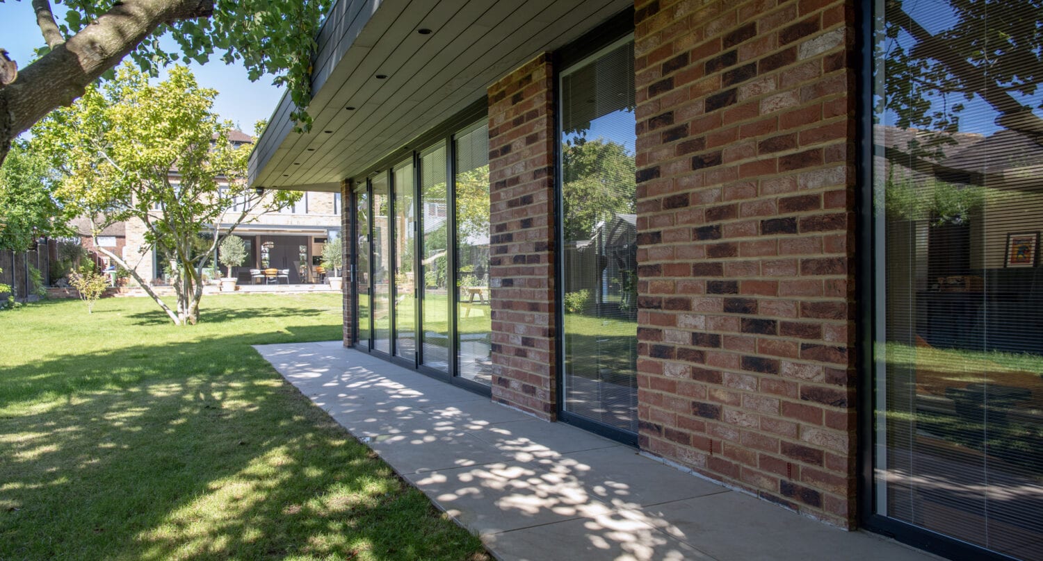 A modern brick house with large glass windows and bifold doors overlooks a sunny backyard with green grass, trees, and a patio area in the distance. Shadows from the trees create patterns on the walkway.