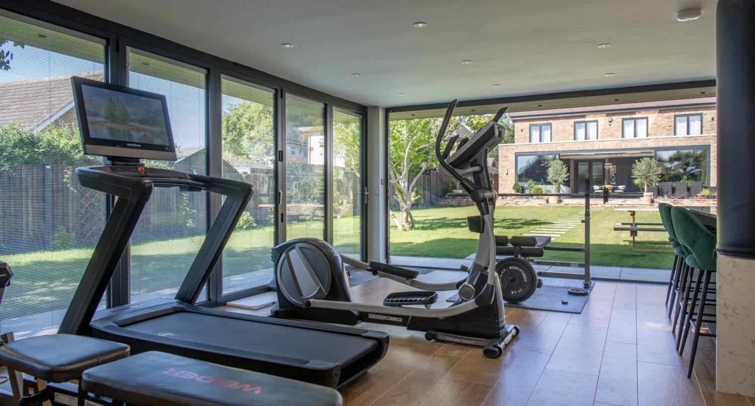 A modern home gym with large windows and sliding doors features a treadmill, elliptical, stationary bike, and weights. The room overlooks a green garden and a large house in the background.