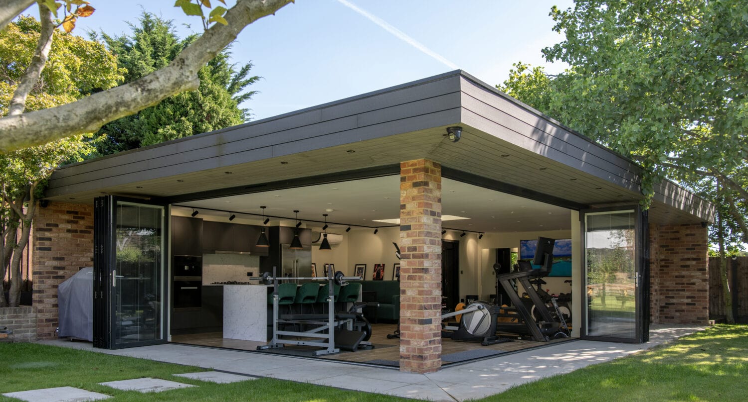Modern garden room with brick pillars and large bespoke glass solutions, featuring open sliding doors that reveal a home gym with exercise equipment and green chairs inside, surrounded by lush trees and a neatly mowed lawn.