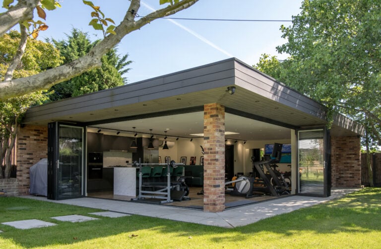 Modern garden room with brick pillars and large bespoke glass solutions, featuring open sliding doors that reveal a home gym with exercise equipment and green chairs inside, surrounded by lush trees and a neatly mowed lawn.