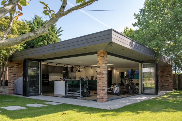 Modern garden room with brick pillars and large bespoke glass solutions, featuring open sliding doors that reveal a home gym with exercise equipment and green chairs inside, surrounded by lush trees and a neatly mowed lawn.