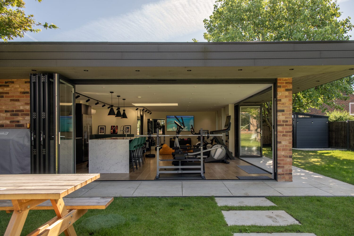 A modern home gym with large sliding doors opens to a sunny garden. Inside, gym equipment is visible, and a marble counter divides the space. Outside on the grass sits a wooden picnic table.