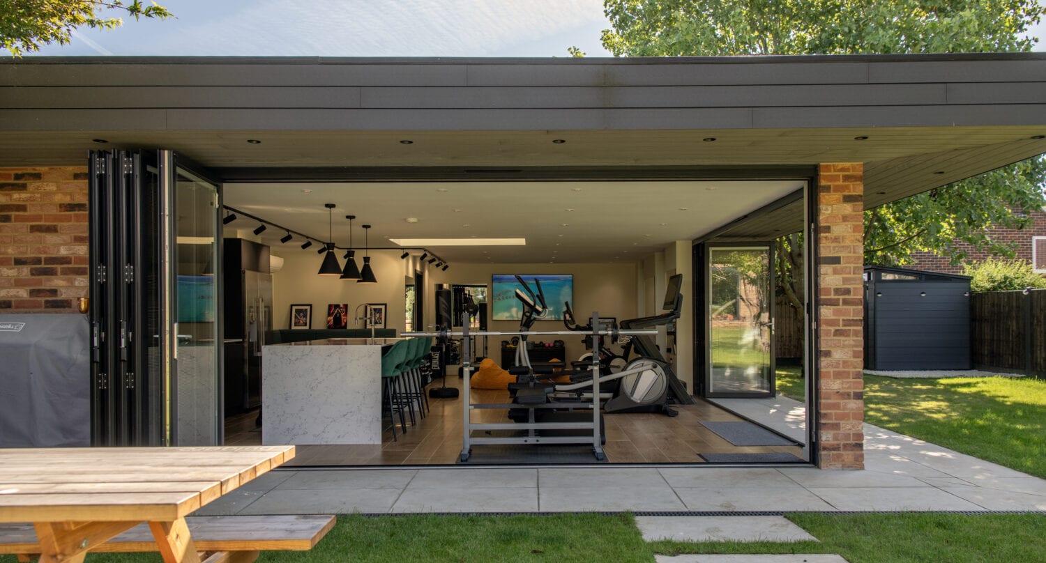 A modern home gym with large sliding doors opens to a sunny garden. Inside, gym equipment is visible, and a marble counter divides the space. Outside on the grass sits a wooden picnic table.