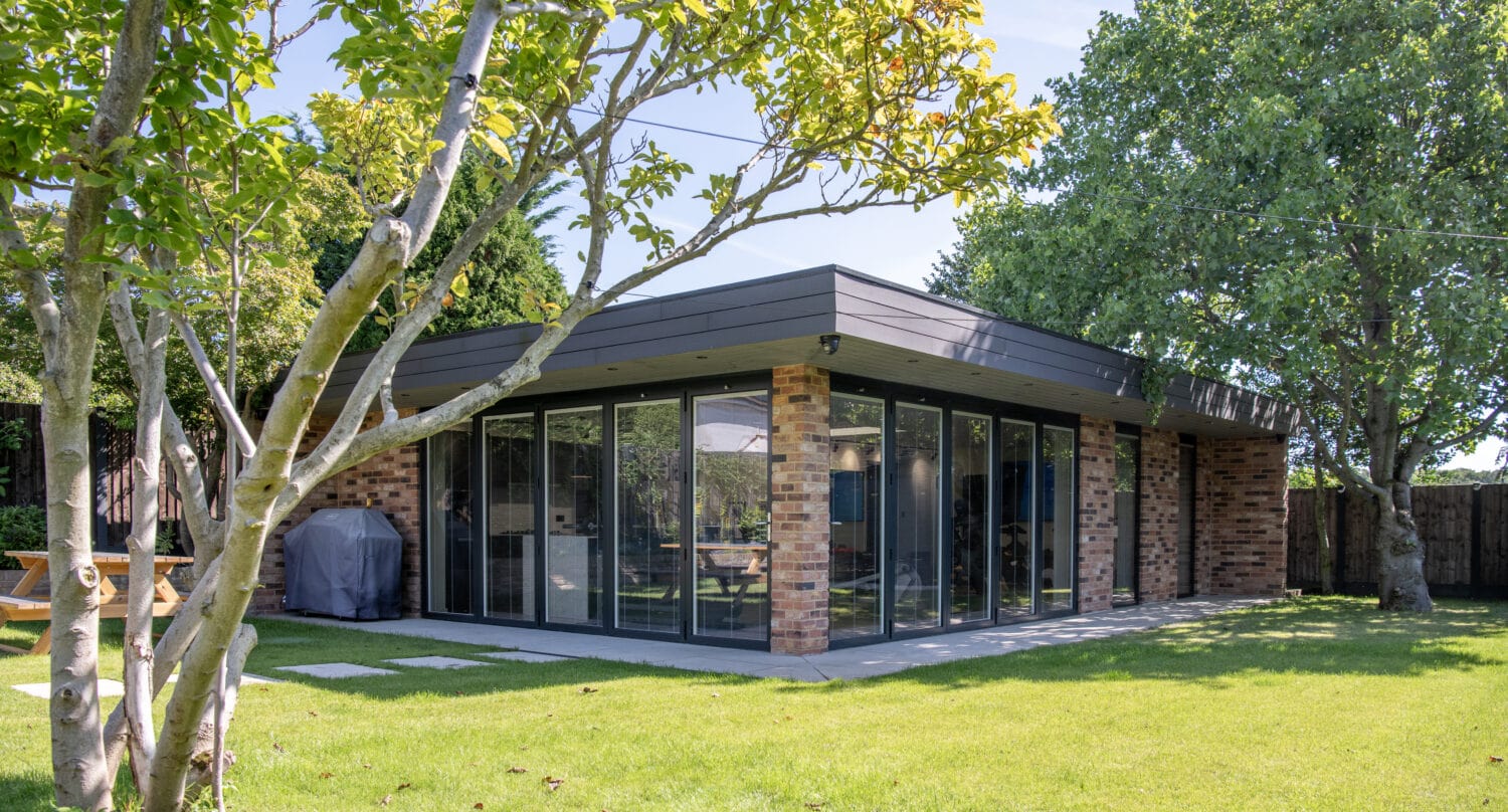 Modern brick and glass house with large floor-to-ceiling windows and sleek sliding doors, surrounded by lush green grass, trees, and a tidy garden on a sunny day.