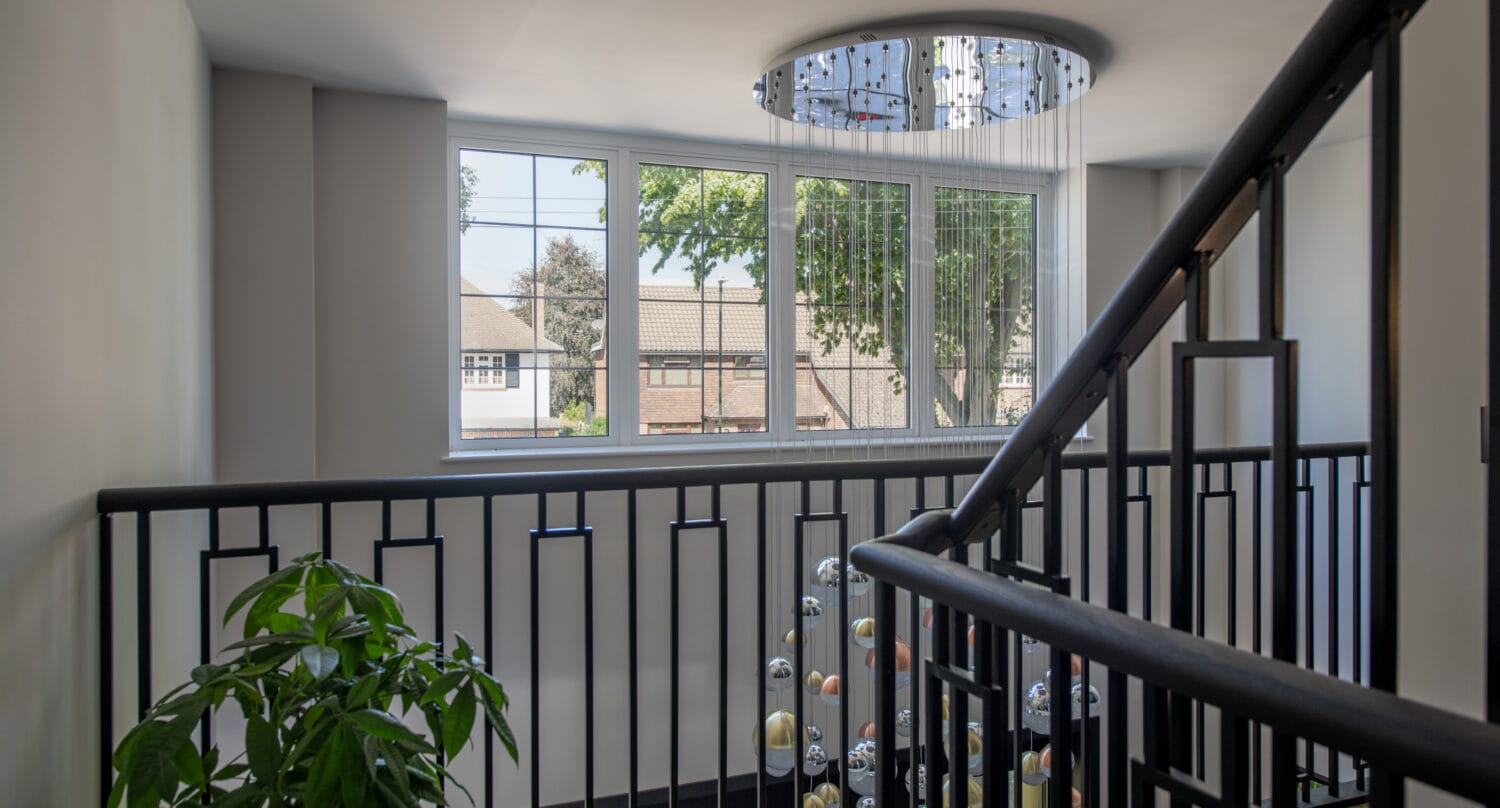 A modern staircase landing with sleek black metal railings, a large window overlooking houses and trees, a green leafy plant, and a contemporary chandelier—enhanced by bespoke glass solutions for added style and light.