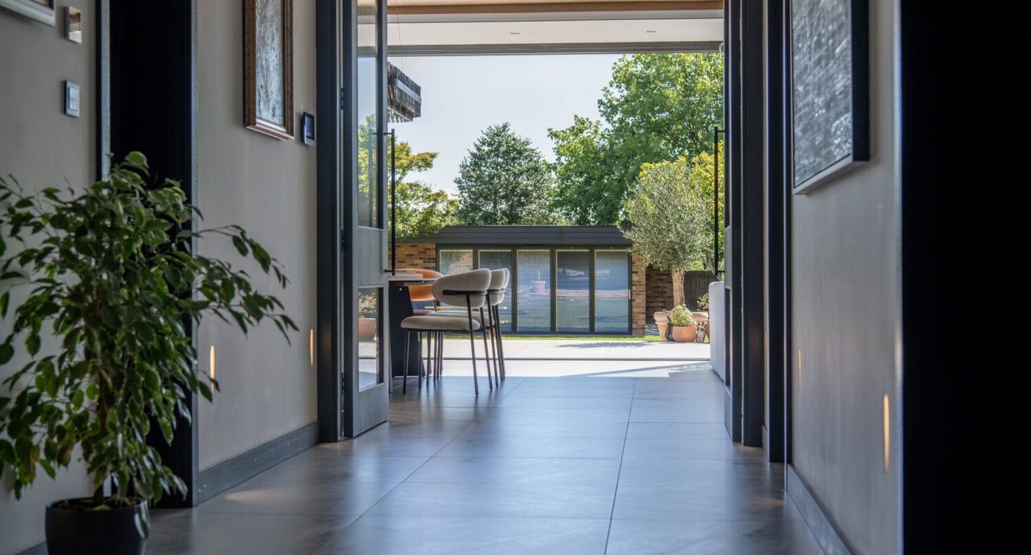 A bright hallway leads to an open dining area with two chairs and a table, overlooking a sunny backyard with green trees and a modern garden shed. Sliding doors frame the view, and a potted plant sits on the left side of the hallway.