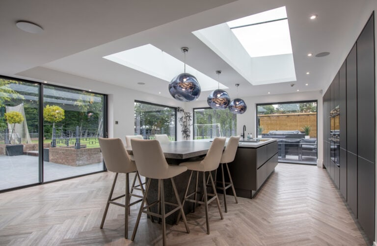 Modern kitchen with large skylights, an island with four high chairs, pendant lights, and floor-to-ceiling bespoke glass solutions including sliding doors opening to a patio and garden, allowing natural light to fill the space.