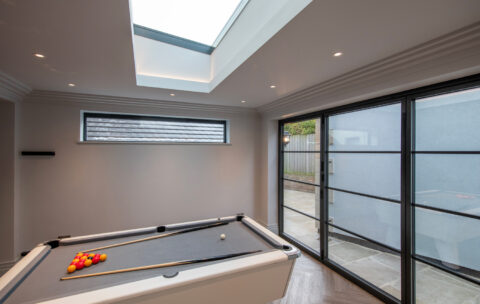 A modern room with a skylight, bifold doors, and a white pool table set up with balls, including a white cue ball and a triangle of red and yellow balls. The space features gray walls and dark wood flooring for a stylish look.