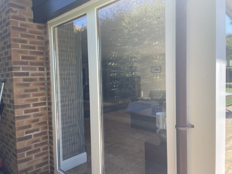 A glass sliding door, part of bespoke glass solutions, sits beside a brick wall reflecting trees and sunlight. Inside, a sofa, coffee table, and tiled floor are visible, with greenery seen through another window in the background.