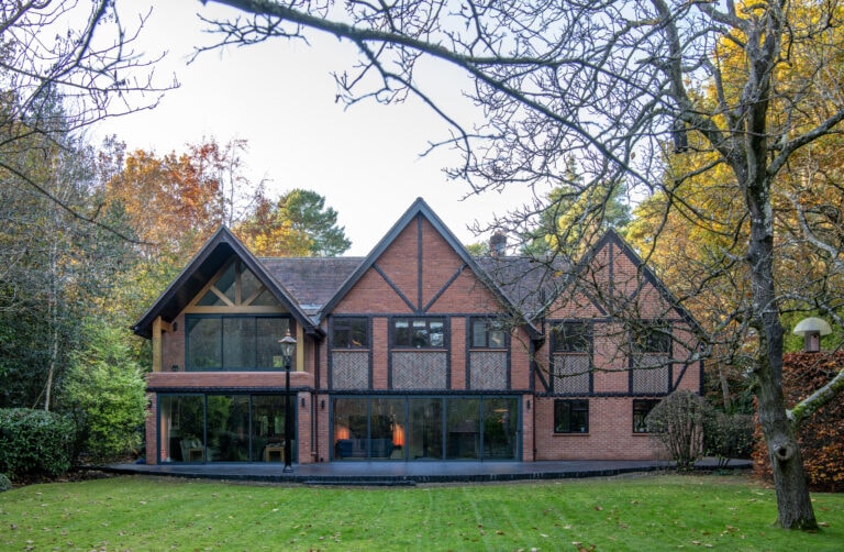 A large modern two-story brick house with bespoke glass solutions and expansive sliding doors, surrounded by trees with autumn foliage and a well-kept green lawn in the foreground.